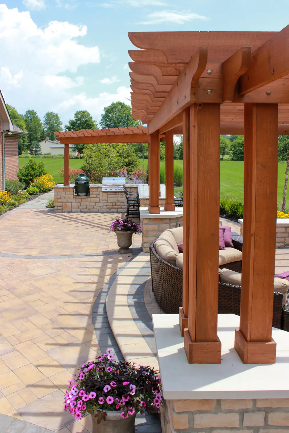 Wood pergola columns, stone column bases, paver patio, grill counter, lounge seating, and container flowers.